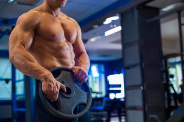 Sportif formda, kaslı, beyaz bir adam elinde ağırlık plakası tutuyordu. Çağdaş spor salonunda siyah şortlu, bulanık, mavi bir arka planı vardı.