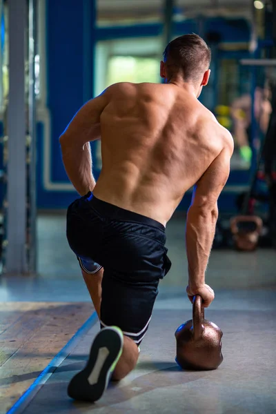 stock image Athletic fit sporty muscular caucasian man doing pulls with brown iron kettlebell in contemporary gym topless in black shorts and lime green sneakers, blurred bluish background