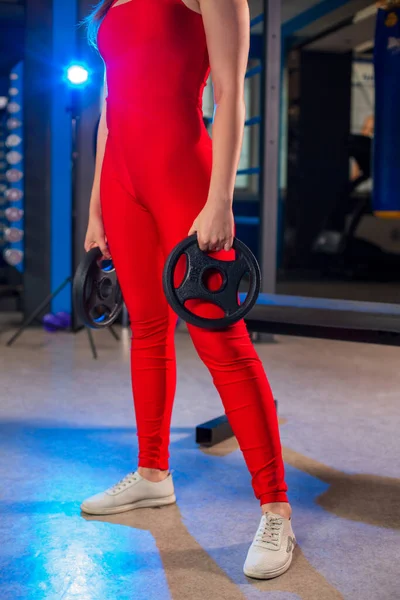 アスレチックフィットスポーティなスリムCaucasian女の子保持黒重量ディスクプレートで彼女の手で赤ジャンプスーツで現代ジム ぼやけた青の背景 — ストック写真