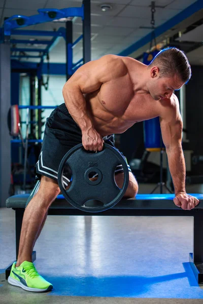 stock image Athletic fit sporty muscular caucasian man holding weight disc plate in his hands while doing pulls on a bench, topless in black shorts in contemporary gym, blurred bluish background