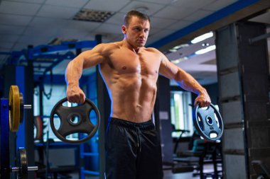 Sportif formda, kaslı beyaz bir adam elinde iki ağırlık plakası tutarken çağdaş spor salonunda siyah şortla omuz sallarken bulanık mavi arka plan.