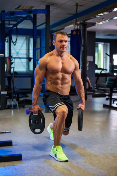 stock image Athletic fit sporty muscular caucasian man holding two weight disc plates in his hands while doing lunges topless in black shorts in contemporary gym, blurred bluish background