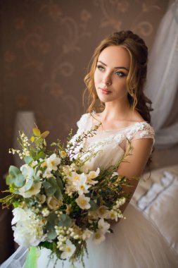 Modaya uygun beyaz elbisesi içinde, elinde bir buket yeşil ve beyaz çiçekle otel odasının penceresinin yanında duran güzel, genç, ince, seksi beyaz bir gelinin orta boy portresi.