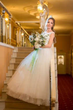 Elinde bir buket yeşil ve beyaz çiçekle otel odasının merdivenlerinde duran zarif, ince, seksi, beyaz gelinlik giymiş beyaz bir kadının portresi.