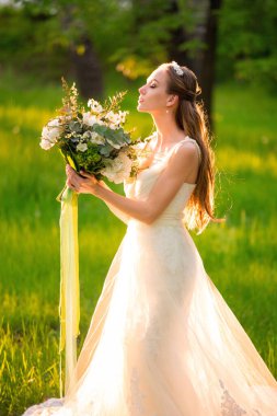 Süslü, genç, ince, beyaz bir gelinin orta boy portresi. Moda modasına uygun beyaz bir elbise içinde. Elinde çiçek buketi var.