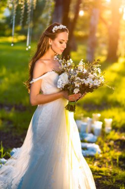 Süslü, genç, ince, beyaz bir gelinin orta boy portresi. Moda modasına uygun beyaz bir elbise içinde. Elinde çiçek buketi var.