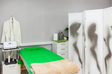 Parlak, modern masaj odasının iç detayları, bej battaniyeli yeşil masaj masası, duvarda kadın silueti ve banyo bornozu resmi olan bölme ekranı.