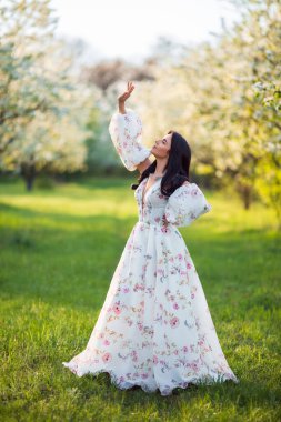 Güzel esmer beyaz bir kadının portresi. Çiçek açan elma bahçesinde beyaz çiçekli bir elbiseyle duruyor. Taze yeşil bir çimenin üzerinde.