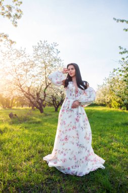 Güzel esmer, beyaz bir kadının portresi. Çiçekli bir elma bahçesinde, yeşil çimlerin üzerinde beyaz çiçekli bir elbiseyle duruyor ve tek eliyle saçına dokunuyor.