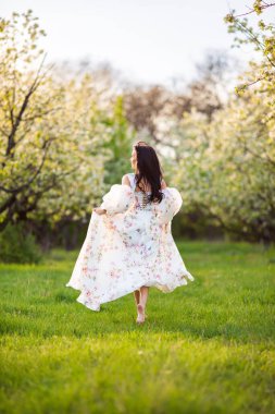 Beyaz çiçekli bir çiçekli elbiseyle koşan esmer, beyaz bir kadının uzun boylu portresi. Sırtından taze yeşil çimenler fırlamış, saçları uçuşuyor.