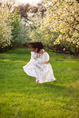Beyaz çiçekli bir çiçekli elbiseyle koşan esmer, beyaz bir kadının uzun boylu portresi. Sırtından taze yeşil çimenler fırlamış, saçları uçuşuyor.