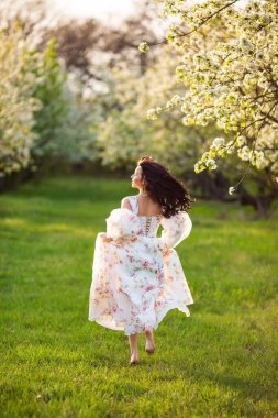 Beyaz çiçekli bir çiçekli elbiseyle koşan esmer, beyaz bir kadının uzun boylu portresi. Sırtından taze yeşil çimenler fırlamış, saçları uçuşuyor.