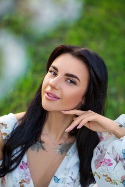 Güzel esmer beyaz bir kadının portresi. Çiçek açan elma bahçesinde beyaz çiçekli bir elbiseyle oturuyor. Yüzüne dokunan yeşil çimenlerin üzerinde.