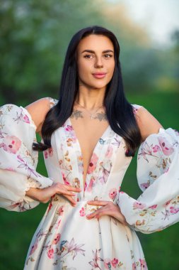 Güzel esmer beyaz bir kadının orta boy portresi çiçek açan elma bahçesinde taze yeşil çimlerin üzerinde beyaz çiçekli bir elbiseyle duruyor.