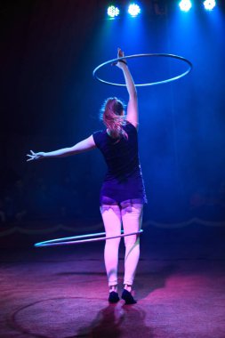 Genç, zayıf, çekici sirk jimnastikçisi kadın, kırmızı arenada hula hoopla canlı performans sergiliyor. Seyirciler heyecanlanıyor.