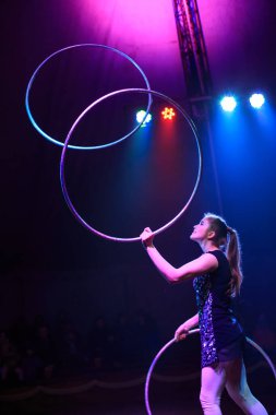 Genç, zayıf, çekici sirk jimnastikçisi kadın, kırmızı arenada hula hoopla canlı performans sergiliyor. Seyirciler heyecanlanıyor.