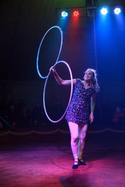 Genç, zayıf, çekici sirk jimnastikçisi kadın, kırmızı arenada hula hoopla canlı performans sergiliyor. Seyirciler heyecanlanıyor.