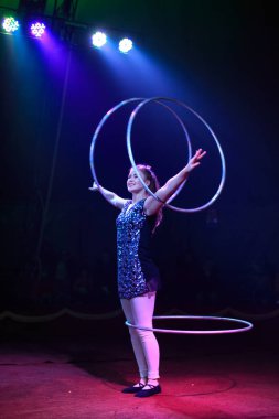 Genç, zayıf, çekici sirk jimnastikçisi kadın, kırmızı arenada hula hoopla canlı performans sergiliyor. Seyirciler heyecanlanıyor.