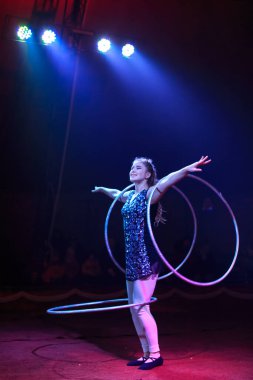 Genç, zayıf, çekici sirk jimnastikçisi kadın, kırmızı arenada hula hoopla canlı performans sergiliyor. Seyirciler heyecanlanıyor.