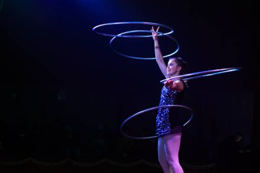 Genç, zayıf, çekici sirk jimnastikçisi kadın, kırmızı arenada hula hoopla canlı performans sergiliyor. Seyirciler heyecanlanıyor.