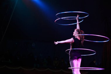 Genç, zayıf, çekici sirk jimnastikçisi kadın, kırmızı arenada hula hoopla canlı performans sergiliyor. Seyirciler heyecanlanıyor.