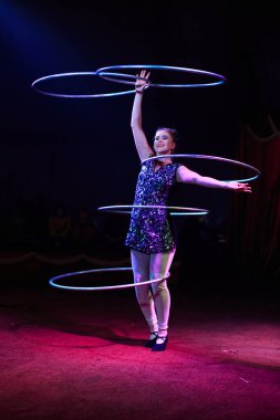 Genç, zayıf, çekici sirk jimnastikçisi kadın, kırmızı arenada hula hoopla canlı performans sergiliyor. Seyirciler heyecanlanıyor.