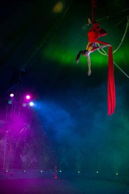 Profesyonel hava kayışları ipek kadın jimnastikçi sirk performansı mavi arenada arka planda renkli konser ışıklarıyla, jimnastik elementlerini sergiliyor.