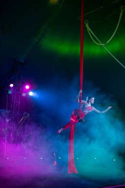Profesyonel hava kayışları ipek kadın jimnastikçi sirk performansı mavi arenada arka planda renkli konser ışıklarıyla, jimnastik elementlerini sergiliyor.