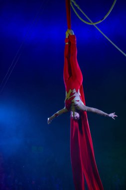 Profesyonel hava kayışları ipek kadın jimnastikçi sirk performansı mavi arenada arka planda renkli konser ışıklarıyla, jimnastik elementlerini sergiliyor.