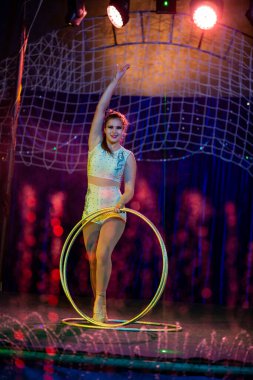 Genç, zayıf, çekici sirk jimnastikçisi kadın, kırmızı arenada hula hoopla canlı performans sergiliyor. Seyirciler heyecanlanıyor.