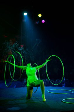 Genç, zayıf, çekici sirk jimnastikçisi kadın, kırmızı arenada hula hoopla canlı performans sergiliyor. Seyirciler heyecanlanıyor.