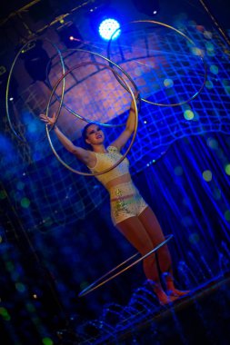 Genç, zayıf, çekici sirk jimnastikçisi kadın, kırmızı arenada hula hoopla canlı performans sergiliyor. Seyirciler heyecanlanıyor.