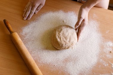 Elleri un kaplı hamurun üzerinde oklavayla çalışıyor. Görüntü, ahşap mutfak yüzeyindeki geleneksel pişirme sürecini yakalıyor ve kırsal bir ev yemeği deneyimini vurguluyor.