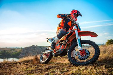 Motokros sporcusu, parlak güneşli bir günde kayalık bir yolda hızlandırıyor, koruyucu giysiler ve turuncu kıyafetler giyiyor. Off-road macera ve ekstrem sporlar