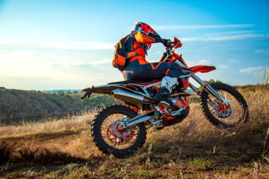 Turuncu ve siyah kıyafetli off-road motosikletçisi gün batımında çimenli bir tepeye tırmanıyor. Dinamik aksiyon ve açık hava sporları konsepti