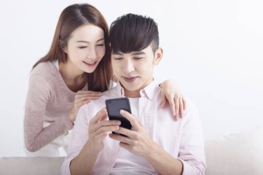 Mutlu genç Asyalı çift cep telefonu kullanıyor, internete takılıyor, internette sohbet ediyor, evdeki kanepede dinleniyor.