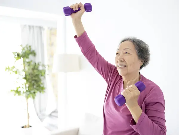 Asian senior woman doing exercises with dumbbells indoors.