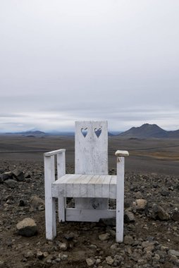 Kalp kesikleri olan beyaz bir sandalye bulutlu bir gökyüzünün altındaki kayalık bir arazide oturuyor..