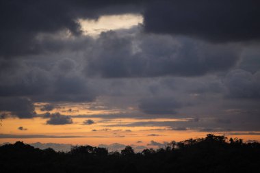 Kosta Rika 'da gün batımı: Siluetli bir ağaç hattının üzerinde, turuncu gökyüzünün içinden baktığı kara bulutlar.