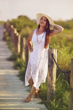 Beyaz elbiseli ve güneş şapkalı güzel bir kadın sahile giden yolda duruyor.