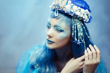 Mavi makyajlı ve deniz kabuğu tacı takan otantik görünümlü bir deniz kraliçesi. Fantezi ve okyanus teması için mükemmel güçlü ve dramatik görüntü