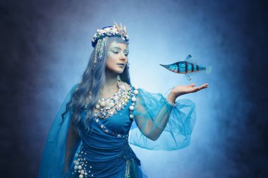 Elinde balık olan kaprisli bir denizkızı. Mavi fantezi, deniz kabuğu tacı ve ruhani güzellik. Denizin büyüsünü yakala.