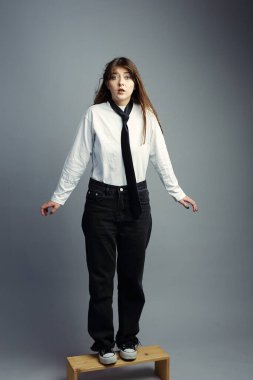 Gömlek ve kravat takan genç bir kadın, küçük bir taburede dururken şok olmuş bir ifadeye sahiptir. Sürpriz konsepti, hazırlıksız yakalanmak, işyeri endişesi ve yeni bir rolde incinebilirlik..