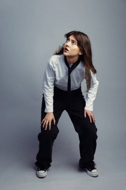 Beyaz gömlekli ve kravatlı ürkmüş genç bir kadın ellerini dizlerinin üstüne çöküp geniş gözlerle yukarı bakıyor. Saçları hafif dalgalı. Tam boy stüdyo çekimi, gri arkaplan.