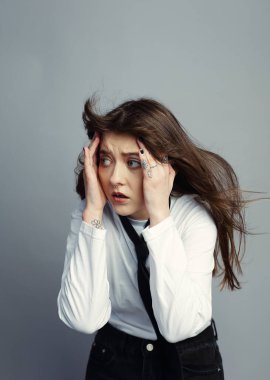 Saçlarını dalgalandıran stresli genç bir kadın başını tutuyor, korkunç bir şekilde yana bakıyor. Beyaz gömlek ve kravat. Gri stüdyo geçmişi.