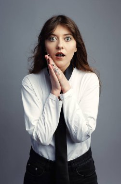 Geniş gözlü ve ağzı açık genç bir kadın şokta ya da şaşkınlık içinde elleri yüzüne yapışık bir şekilde soluk soluğa kalıyor. Şık bir şekilde beyaz gömlek ve kravat giymişti. Gri stüdyo geçmişi.