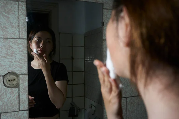 漂亮的女人在浴室的镜子前用剃须泡沫擦拭她的脸 — 图库照片