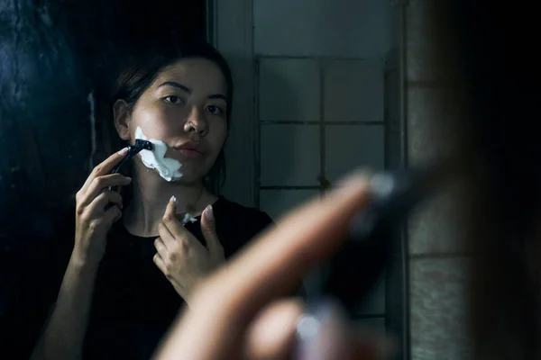 女人手里拿着剃须刀 站在浴室的镜子前 把剃须泡沫涂在下巴上 — 图库照片