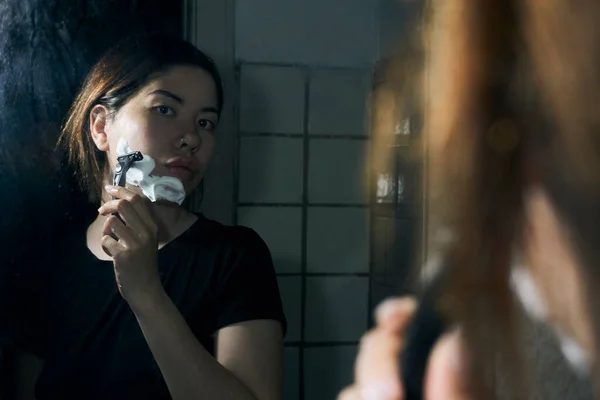 女人手里拿着剃须刀 站在浴室的镜子前 把剃须泡沫涂在下巴上 — 图库照片