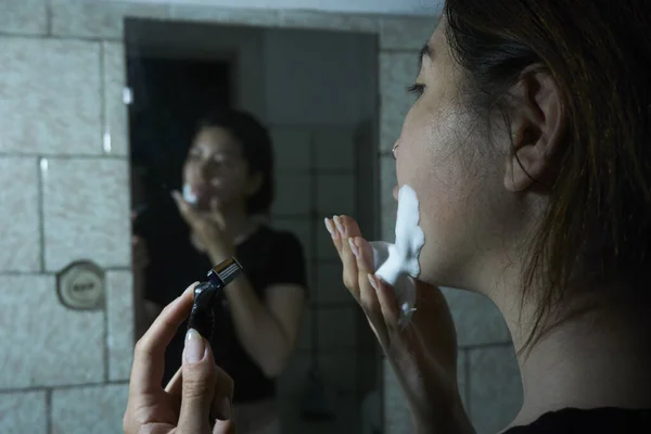 女性は手にカミソリを持ちバスルームミラーに立っている間に顎にシェービングフォームを適用します ストック画像